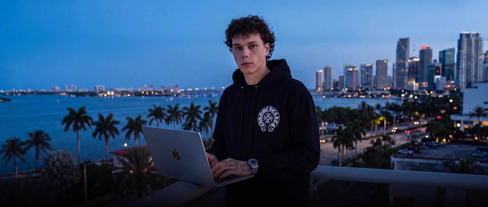 Brez in his Chrome Hearts hoodie working on his laptop on a Miami rooftop at blue hour